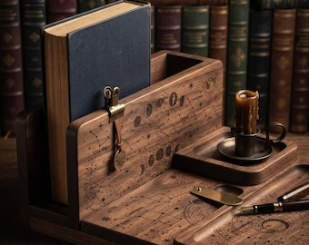 May include: A dark wood desk organiser featuring engraved celestial patterns, including constellations and moon phases. It holds a dark blue book, a brass clip with a small circular charm, a small candle in a metal holder, and a fountain pen.