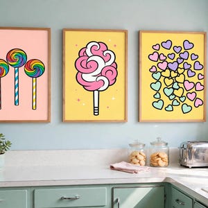 May include: Three framed wall art prints featuring colorful illustrations. The left print shows three rainbow lollipops on a pink background. The center print displays pink cotton candy on a yellow background. The right print features many small pastel hearts on a yellow background.