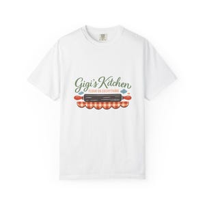 Peut inclure: Un t-shirt blanc à manches courtes avec une impression graphique portant le texte Gigi's Kitchen et FLOUR ON EVERYTHING. Le motif comprend un rouleau à pâtisserie gris foncé aux poignées rouges au-dessus d'une bordure festonnée à carreaux orange et blancs.