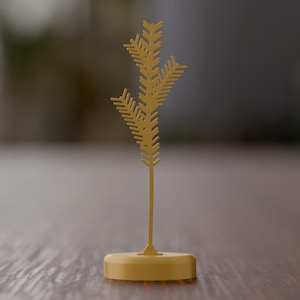 May include: A mustard yellow decorative object featuring a thin, vertical stem topped with a stylized, geometric pine branch silhouette. The entire structure is mounted on a solid, circular base of the same color, creating a singular, upright piece.