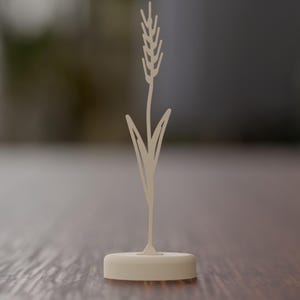 May include: A light beige, flat, two-dimensional cutout of a single stalk of wheat with a pointed head and two long, slender leaves. The wheat stalk is mounted vertically into a solid, oval-shaped, light beige base that sits flat.