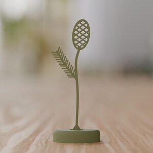 May include: A matte olive green metal sculpture featuring a slender stem rising from a circular base. The stem supports a textured, oval-shaped top with a cross-hatched pattern and a side branch with multiple thin, pointed, leaf-like protrusions.
