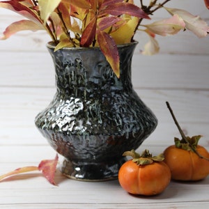 May include: A dark blue ceramic vase with a textured surface holds a bouquet of autumn leaves. Two orange persimmons sit on a white wooden surface next to the vase.