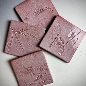 Puede incluir: Cuatro posavasos cuadrados de cerámica con fondo rosa y diseños de hojas blancas prensadas. Los posavasos presentan diferentes patrones de hojas.