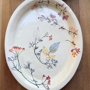 May include: Off-white ceramic oval platter with botanical illustrations. The platter features colorful floral and leaf designs in red, purple, blue, and yellow. The rim is slightly raised, and the overall design is organic and artistic.