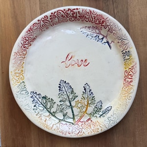 May include: A white ceramic plate with a colorful leaf design and the word "love" in red script. The rim features a decorative pattern with red, yellow, and blue hues. The plate is on a wooden surface.