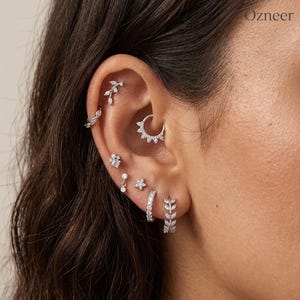 May include: A set of silver-toned earrings featuring a leaf-shaped cartilage stud, a small hoop, a cluster stud, a dangling stone stud, a star stud, and two hoop earrings with leaf and stone accents, all adorned with clear crystals.