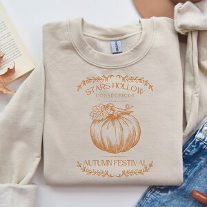 Puede incluir: Una sudadera beige con un diseño gráfico marrón que presenta una calabaza y el texto "Stars Hollow Connecticut Autumn Festival".
