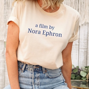 Könnte beinhalten: Ein cremefarbenes T-Shirt mit einem blauen Textdesign, das "a film by Nora Ephron" lautet.