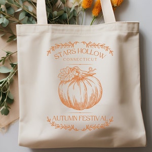 Può includere: Una borsa di tela bianca con un design grafico arancione. Il design presenta una zucca, foglie e il testo "Stars Hollow Connecticut Autumn Festival".