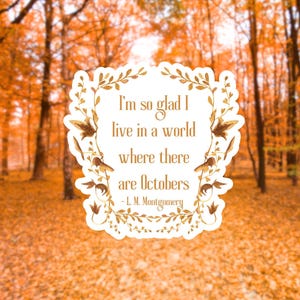 May include: A sticker with the quote "I'm so glad I live in a world where there are Octobers" by L. M. Montgomery, framed by a floral design. The background features a blurred autumn forest with orange and yellow leaves.