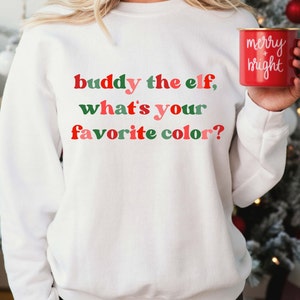 May include: White sweatshirt featuring the phrase "buddy the elf, what's your favorite colour?" in red, green, and pink. A red mug with "merry + bright" in white is held in the hand.