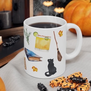 May include: White ceramic mug featuring a black cat, a book, a broom, a cocktail, and autumn leaves. The mug has a black rim and handle.