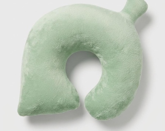 May include: A soft, plush, U-shaped neck pillow in a light sage green color. The pillow features a smooth, velvety texture and a distinct leaf-like shape with a small, rounded stem protruding from the top right side of the cushion.