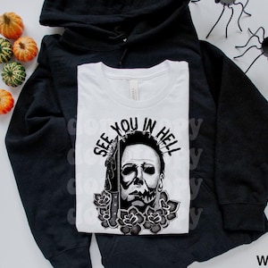 May include: A white t-shirt featuring a black graphic of a masked character holding a knife, surrounded by roses, with the text "SEE YOU IN HELL" arched above. The shirt is layered over a black hooded sweatshirt, accompanied by small decorative pumpkins.