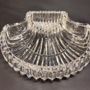 May include: A clear glass dish shaped like a scallop shell, featuring a series of vertical ridges radiating from the center to the scalloped edge. The exterior rim has a faceted, diamond-cut pattern that catches light, creating a reflective surface.