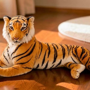 May include: A plush tiger toy featuring a realistic orange and black striped coat, white fur accents on the face and underbelly, and amber-colored eyes. The stuffed animal is depicted in a resting, lying-down position with its head facing forward.