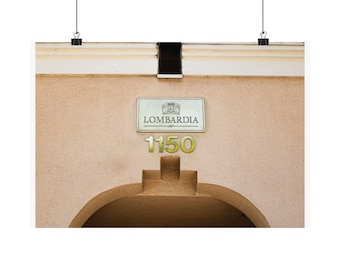 May include: A beige stucco building facade featuring a rectangular sign with the text LOMBARDIA and a crest above it. Below the sign, the metallic house numbers 1150 are mounted above a dark arched entryway. The structure is suspended by two clips.