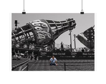 May include: A black and white photograph of a stadium seating area featuring a large Coca-Cola bottle structure overhead. The scene includes rows of empty seats, a person sitting in the middle, and signage reading Made of Cocktail and Ketel One.