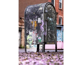 May include: A weathered, dark metal mailbox standing on four legs, heavily covered in layers of graffiti, stickers, and peeling paint. The surface features various illegible markings, tags, and small colorful decals across its curved top and front door.