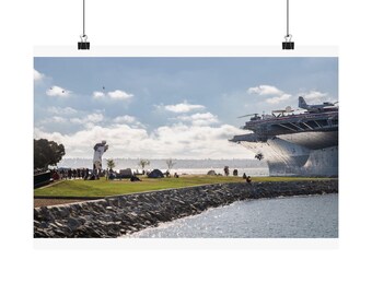 May include: A large outdoor sculpture of a sailor kissing a nurse stands on a grassy waterfront park next to a massive gray aircraft carrier docked in the harbor. The scene includes a rocky shoreline, scattered people, and a bright blue sky.