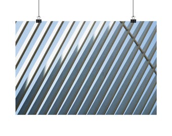 May include: A rectangular wall art print featuring a pattern of diagonal silver metallic slats set against a light blue sky background. The print is suspended by two black metal clips attached to thin black wires at the top edge of the frame.
