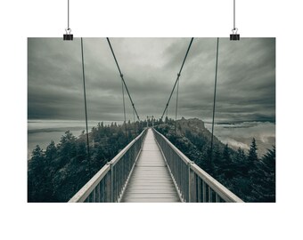 May include: A landscape wall art print featuring a suspension bridge extending into the distance over a mountain ridge. The scene includes dark green pine trees, thick low-hanging clouds, and a moody, overcast gray sky above the rocky terrain.