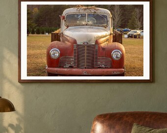 May include: A vintage, weathered truck with a faded red and white exterior, featuring a prominent vertical grille, round headlights, and a worn hood. The vehicle is positioned in an open field with trees in the background, captured in a framed wall art display.