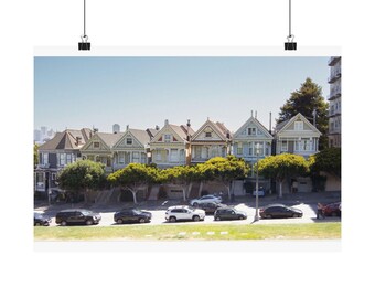 May include: A row of colorful Victorian houses in San Francisco, known as the Painted Ladies, featuring intricate architectural details, gabled roofs, and bay windows, situated above a street with parked cars and green trees under a clear sky.