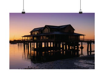 May include: A stilt house stands over calm water at sunset, featuring a dark silhouette against a gradient sky of orange, yellow, and purple. The structure has a wide porch and multiple roof gables, with its reflection visible on the water surface.