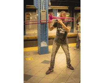 May include: A musician plays a bright magenta trombone while wearing a dark gray t-shirt with the white text JONI NOEL Music, distressed gray jeans, and black and pink sneakers. The scene is set in a subway station with blue structural pillars.