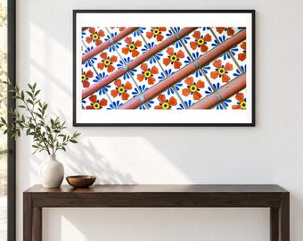 May include: A framed wall art print featuring a repeating pattern of square tiles with orange four-petaled flowers, yellow centers, and blue leaf accents, intersected by diagonal terracotta-colored bars against a white background.