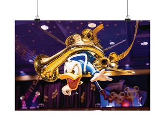 May include: A Donald Duck figure in a blue sailor outfit is suspended in mid-air, surrounded by a large, shiny gold brass instrument. The character has an open beak and is reaching out with one hand, set against a dark purple stage background.