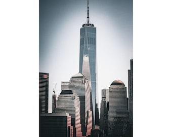 May include: A vertical wall art print featuring the One World Trade Center skyscraper rising above the surrounding New York City skyline. The glass-paneled tower stands tall against a pale sky, with surrounding buildings in shades of gray.