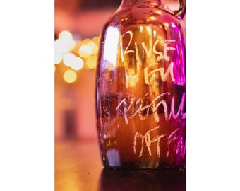 May include: A brown glass growler bottle featuring white etched text that reads RINSE WHEN REFILLED OFTEN. The bottle has a rounded shape with a handle at the top, reflecting warm ambient light with pink and orange hues across its surface.