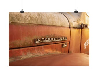 May include: A close-up of a weathered red International K-5 truck hood featuring a metallic emblem with the text INTERNATIONAL and K-5. The paint shows significant signs of oxidation, rust, and fading, with a warm golden light illuminating the surface.