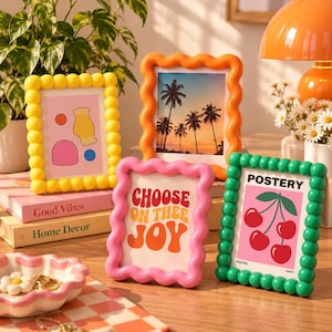May include: Four rectangular picture frames with wavy or beaded borders in yellow, orange, pink, and green. The frames display various prints, including a sunset with palm trees, a cherry illustration with the text POSTERY, and text reading CHOOSE ON THEE JOY.