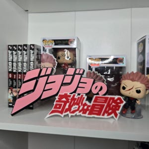 May include: A pink and white sign featuring the Japanese text "ジョジョの奇妙な冒険" positioned in front of several Jujutsu Kaisen manga volumes and two Funko Pop figures in their original boxes, including one character with spiky pink hair and a scar.