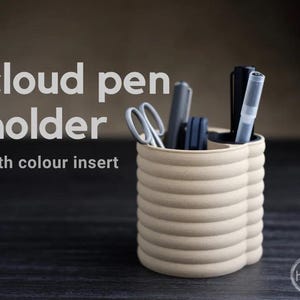 Könnte beinhalten: Ein beigefarbener, wolkenförmiger Stiftehalter mit dunklem Inneneinsatz, der verschiedene Stifte und eine Schere enthält. Der Text "cloud pen holder with colour insert" ist links zu sehen, ein kleines kreisförmiges Logo unten rechts.
