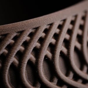Peut inclure: Un objet brun foncé présentant une structure en treillis tridimensionnelle complexe avec des lignes courbes répétitives et des textures en couches visibles. Un petit logo vert circulaire avec le texte h3liø design est situé en bas à droite.