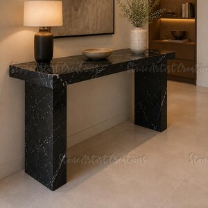 May include: A black marble console table with white veining, featuring a rectangular top and two thick slab legs. On the table sits a black lamp with a white shade, a shallow beige bowl, and a textured white vase containing green foliage.