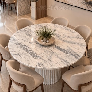 Puede incluir: Una mesa de comedor redonda con tablero de mármol blanco y gris y base cilíndrica estriada, rodeada de seis sillas tapizadas en beige con patas de madera oscura. Un cuenco poco profundo con una planta verde descansa en el centro.
