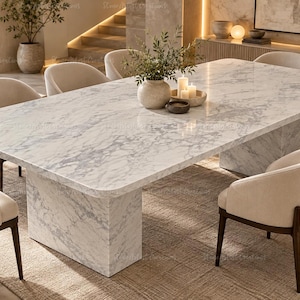 Puede incluir: Una mesa de comedor rectangular con tablero de mármol blanco con vetas grises y base de pedestal a juego. Seis sillas tapizadas en tela color crema con patas de madera oscura están dispuestas alrededor. Un jarrón de cerámica con plantas y velas en el centro.