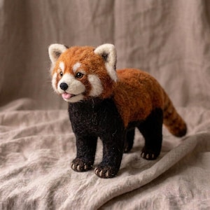 May include: A needle-felted red panda figurine featuring a textured coat of reddish-brown and black wool. The creature has white facial markings, dark eyes, a small black nose, and a tiny pink tongue visible in its open mouth.