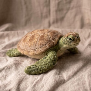 May include: A needle-felted sea turtle figurine featuring a textured brown shell with defined scutes and a green head and flippers. The turtle is depicted in a swimming pose with its front flippers extended outward and its head turned slightly to the side.
