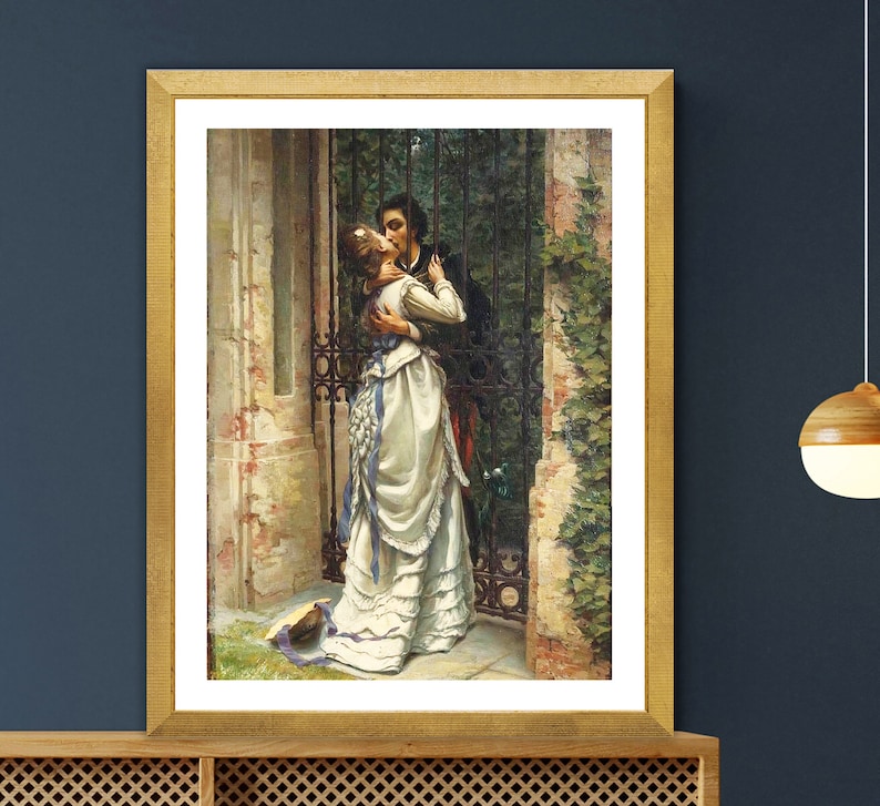 May include: A framed print of a romantic scene. A man and woman embrace in front of a wrought iron gate. The woman wears a white dress with blue accents. The artwork is in a gold frame and is displayed on a wooden cabinet.