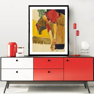 Puede incluir: Una pintura al agua enmarcada de una mujer con un vestido rojo, tumbada sobre un fondo amarillo y marr&oacute;n.