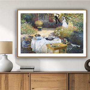 Puede incluir: Una pintura que representa una escena de jard&iacute;n con personas haciendo un picnic. La pintura est&aacute; enmarcada en oro y cuelga de una pared blanca. Hay una l&aacute;mpara y una planta sobre una mesa de madera delante de la pintura.