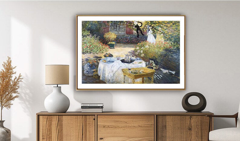 Puede incluir: Una pintura que representa una escena de jard&iacute;n con personas haciendo un picnic. La pintura est&aacute; enmarcada en oro y cuelga de una pared blanca. Hay una l&aacute;mpara y una planta sobre una mesa de madera delante de la pintura.