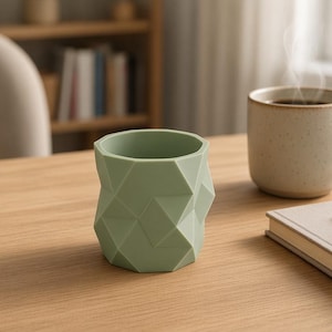 May include: A sage green desk tidy featuring a faceted, geometric exterior with multiple flat triangular and diamond-shaped planes. The container has a wide, circular opening at the top and a solid base, providing a structured, angular appearance.
